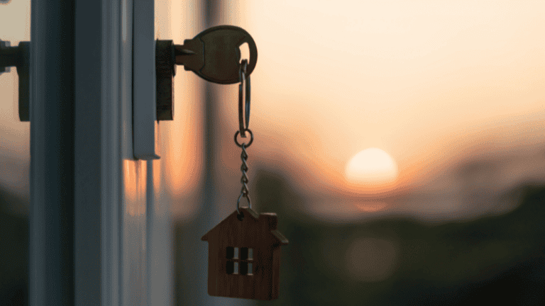 A close-up of a key with a house-shaped keychain in a door lock at sunset, symbolizing home access or ownership.