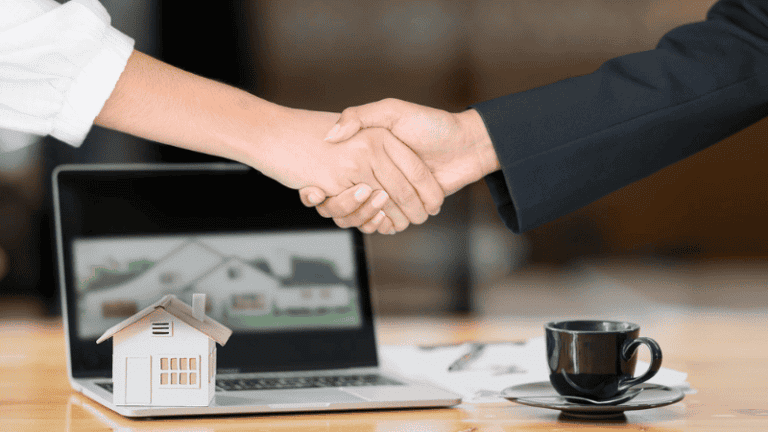 Two people shake hands in front of a laptop displaying a house image, symbolizing a successful property deal, with a miniature house and coffee cup on the table.
