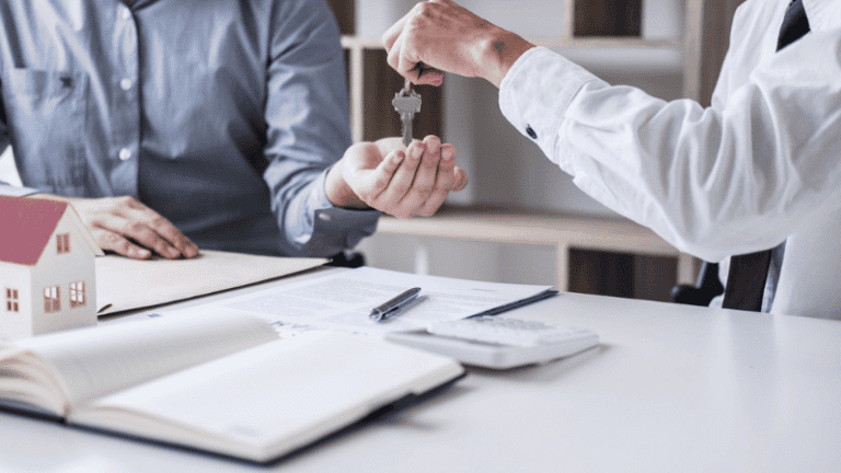 A landlord hands over a key to a tenant during a property agreement meeting, with documents and a small house model on the table.