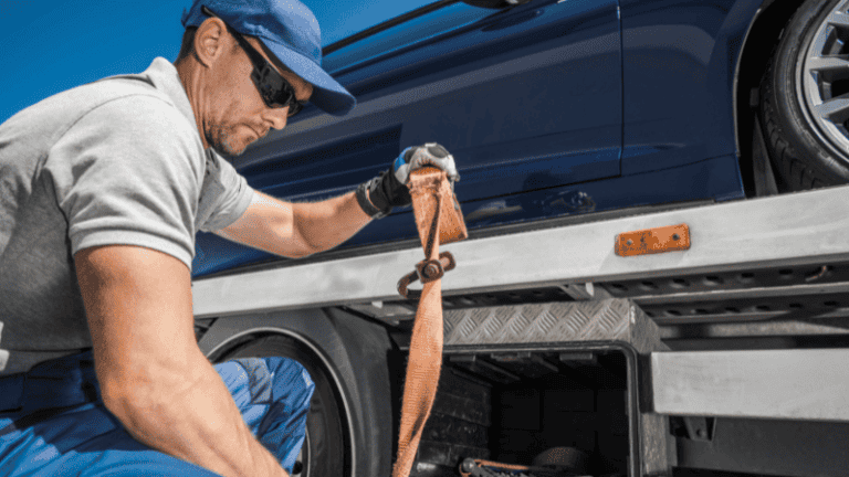 A worker in a blue uniform fastens an orange strap to secure a car on a flatbed tow truck.